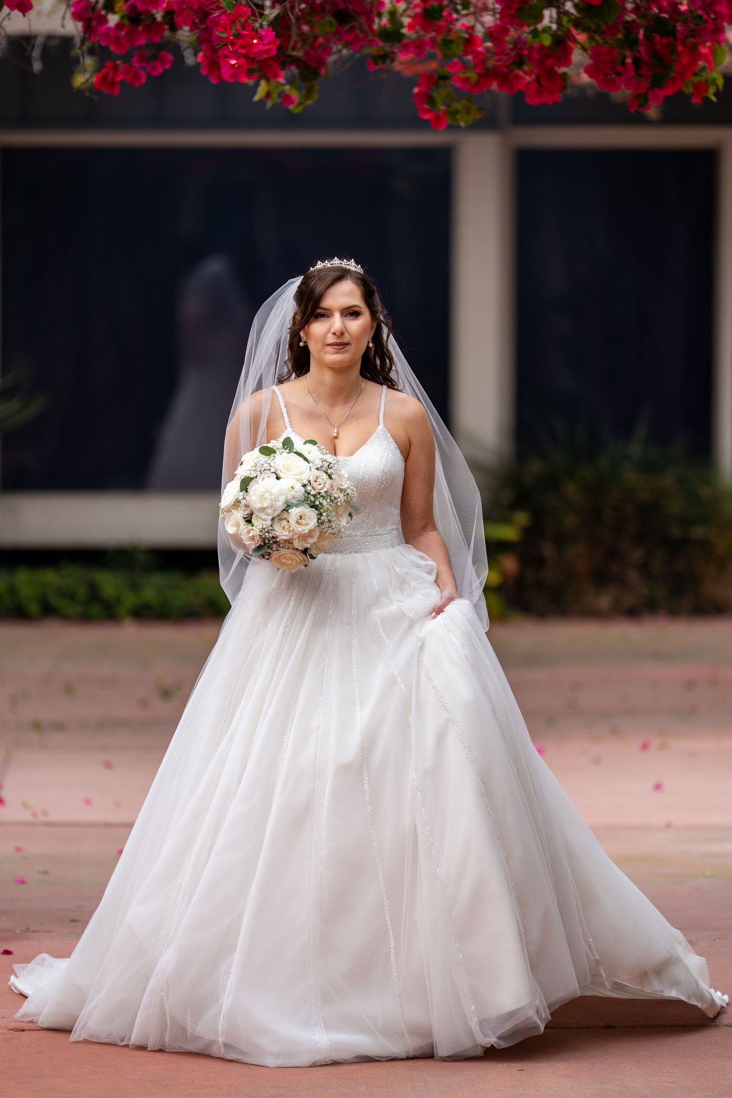Bride in white ball gown with veil and floral bouquet stands outdoors beneath vibrant red flowers.