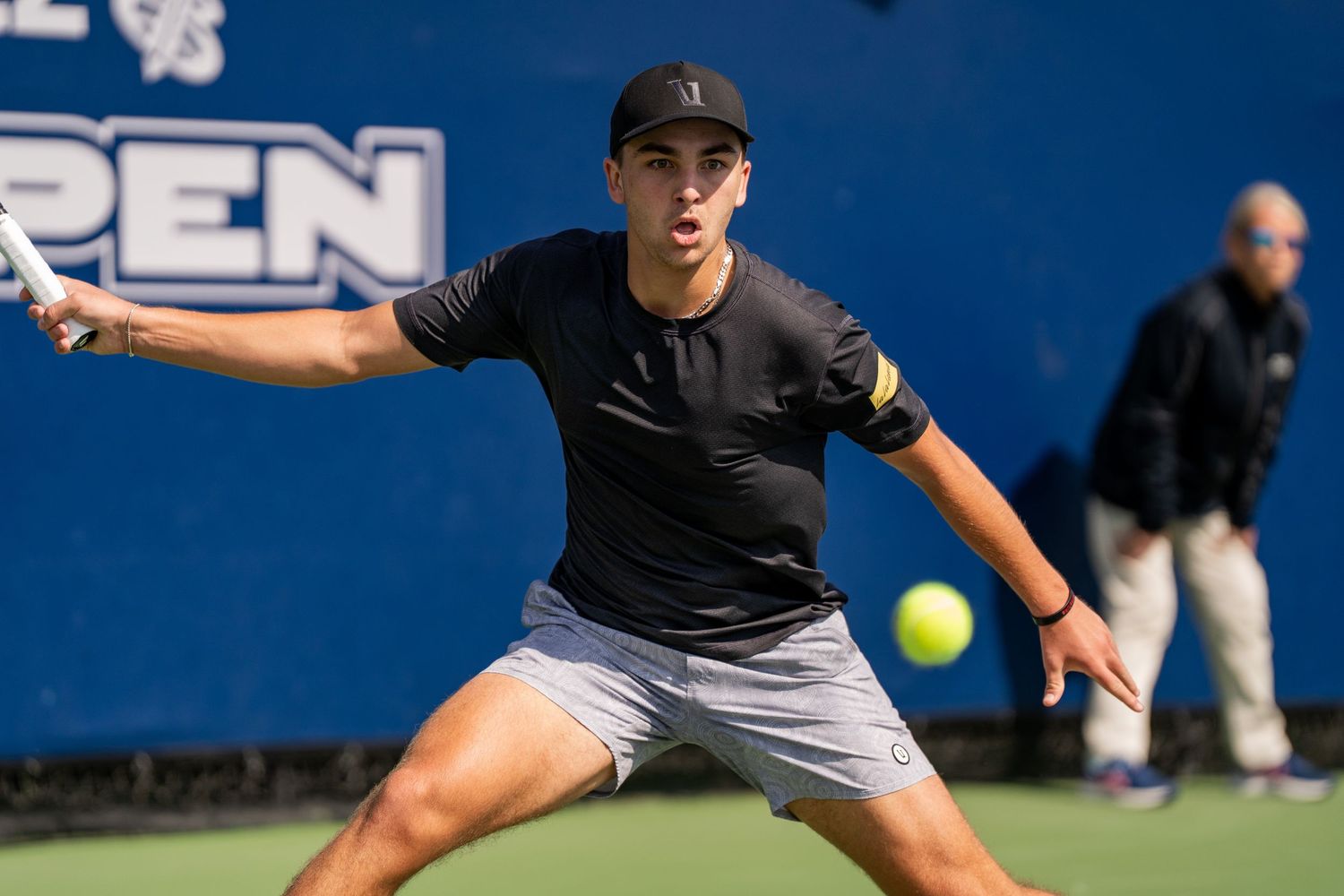 Tennis player in black outfit hitting forehand shot during outdoor tournament match.