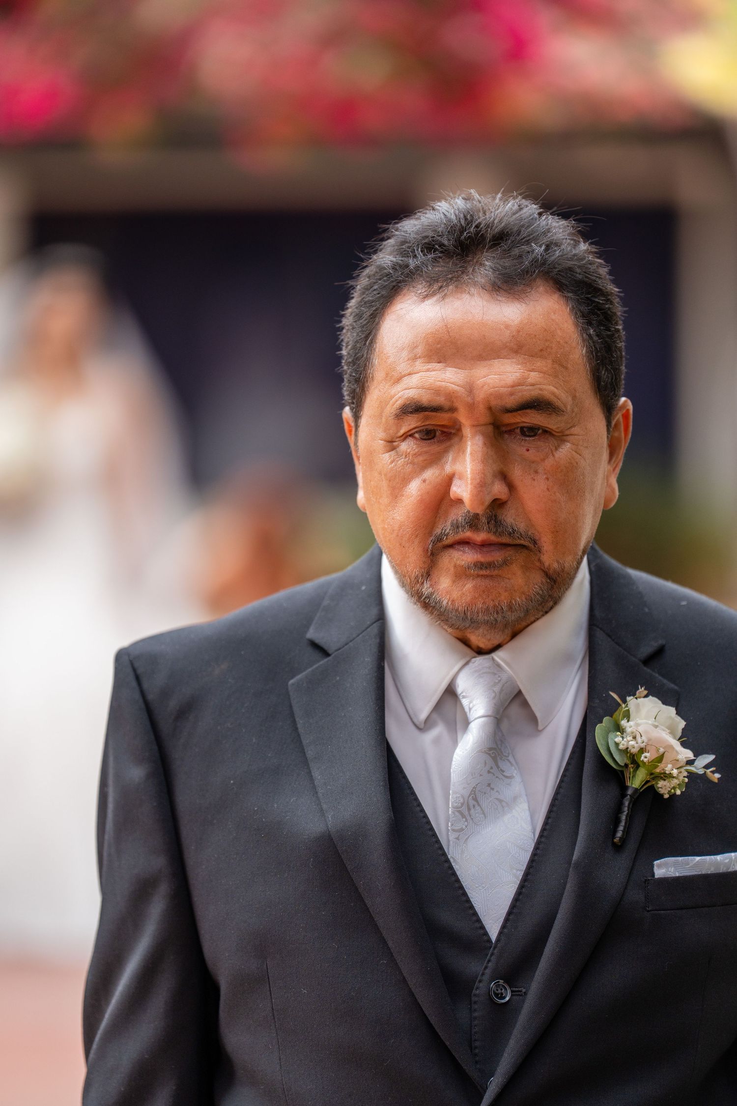 Elegant man in gray suit with boutonniere at wedding, bride visible in background.