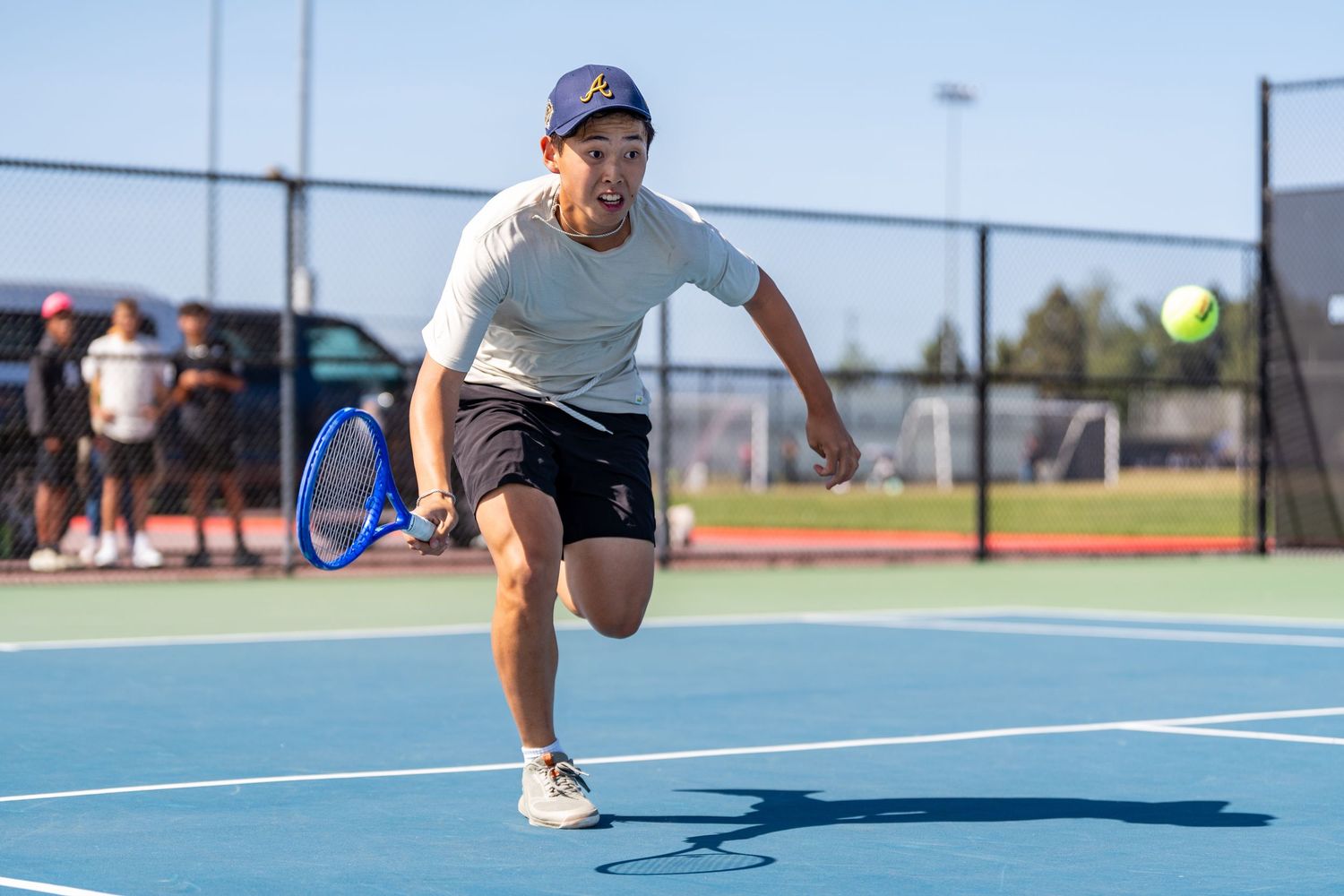 Male tennis player in gray shirt sprints to return a shot on a blue outdoor hard court in sunny weather.