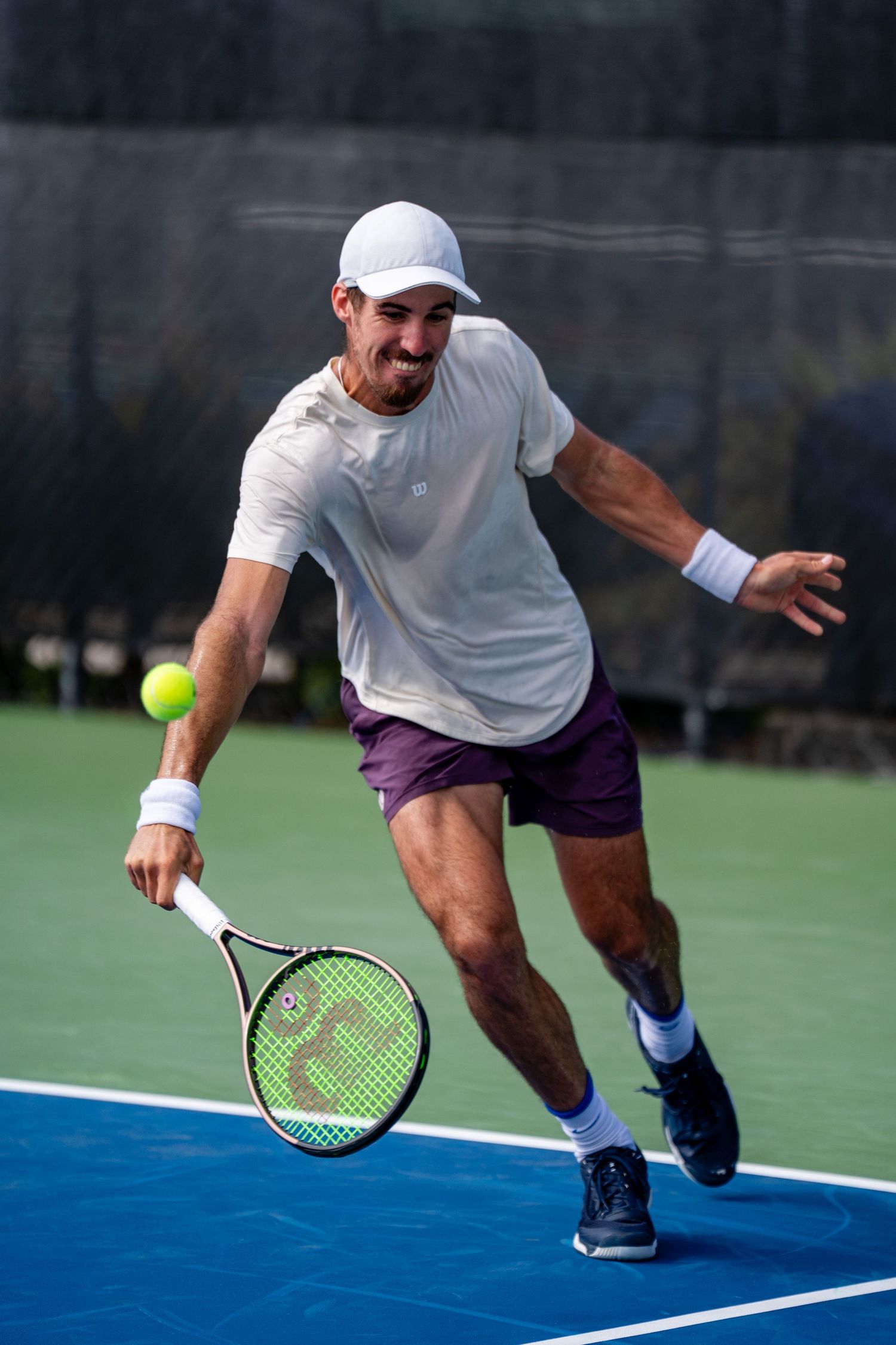 Male tennis player in white cap lunges to hit a forehand on a blue outdoor hard court.