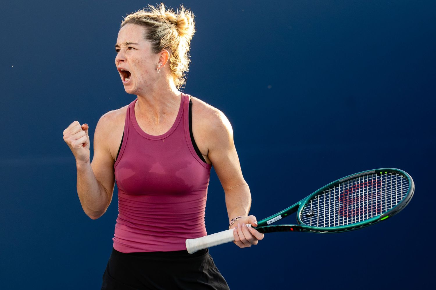 Female tennis player in red top celebrating with fist pump holding Wilson racket on blue court.