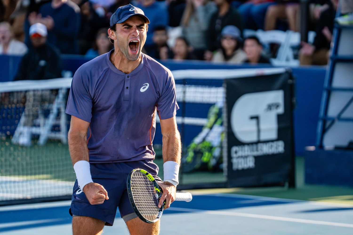 Male tennis player in navy Asics shirt celebrating a point at a Challenger Tour event.