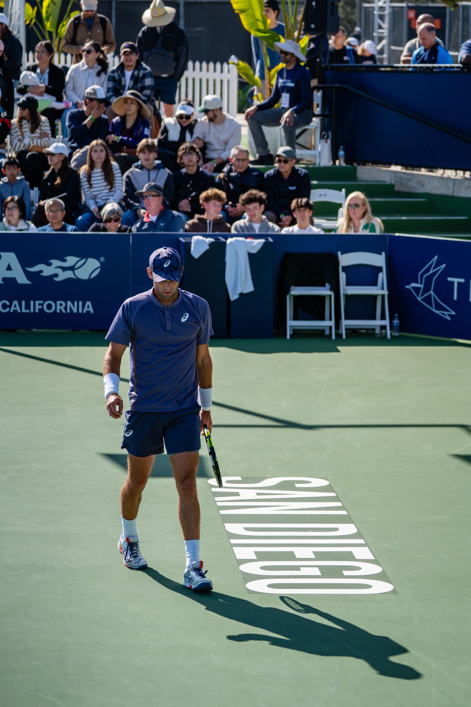 Male tennis player in ASICS gear standing on green court at San Diego Open.