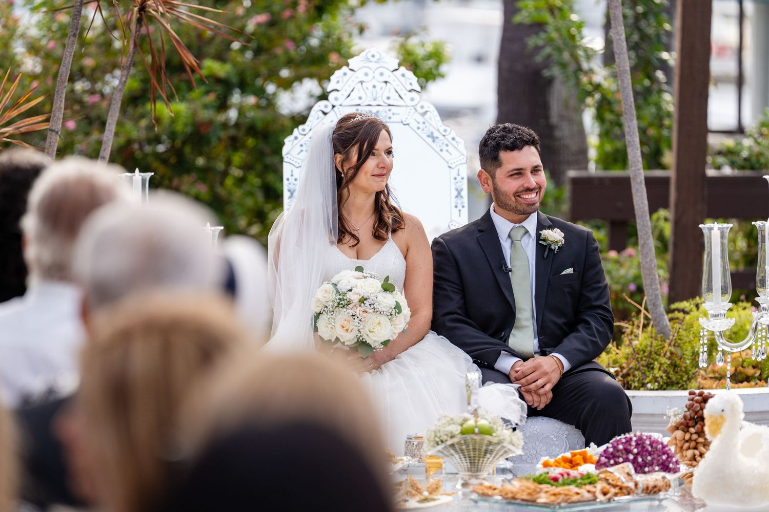 Bride and groom sitting together at outdoor wedding ceremony, bride holding white bouquet, groom in dark suit, elegant decor.