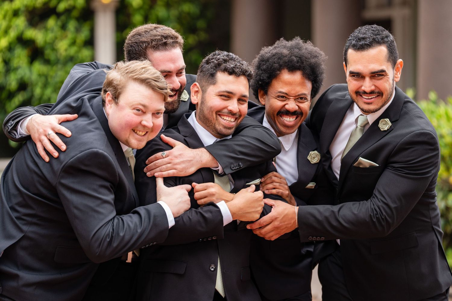 Five groomsmen in dark suits laughing and hugging together outdoors at a wedding celebration.