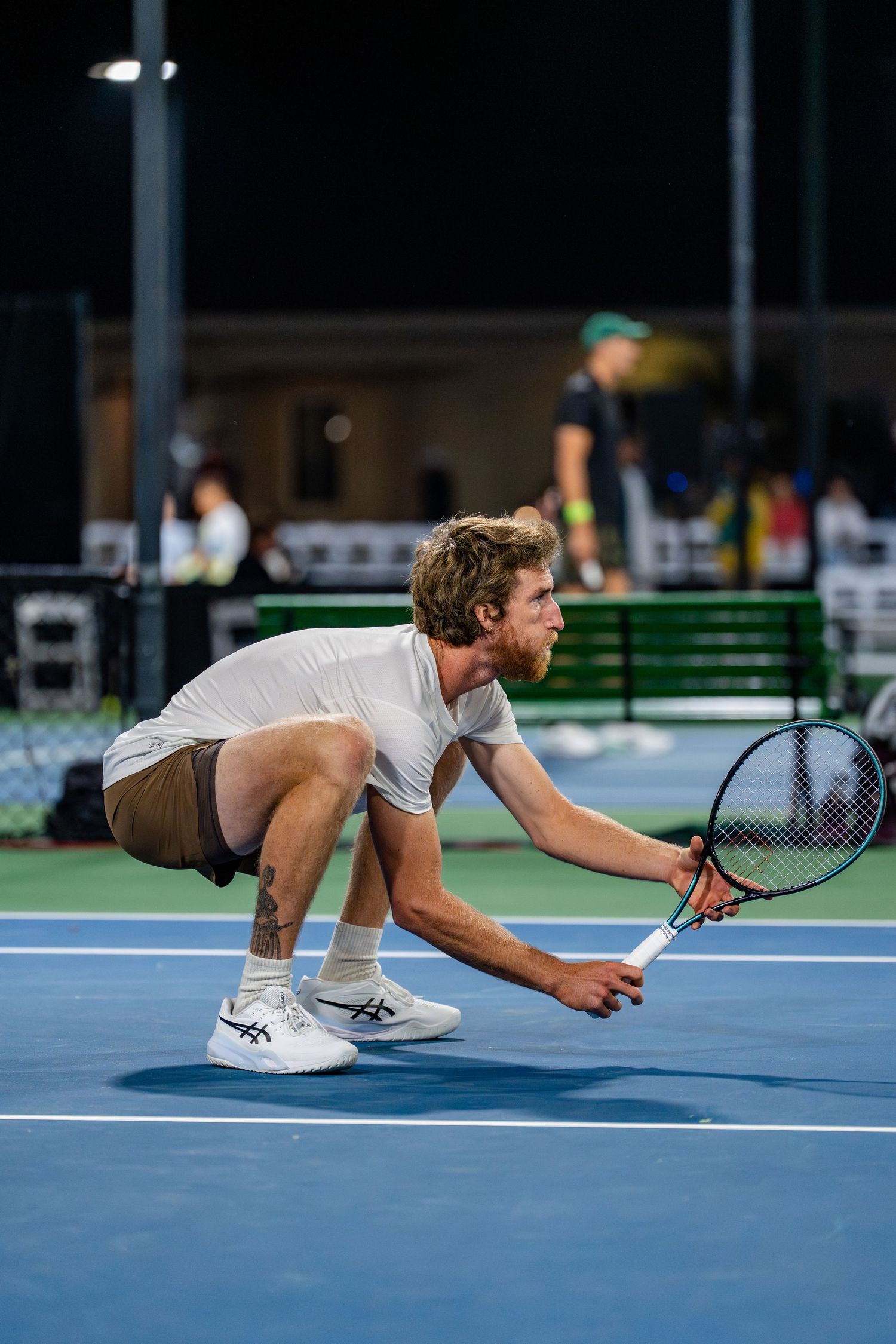 Male tennis player crouches low on blue court at night during an indoor tennis tournament.