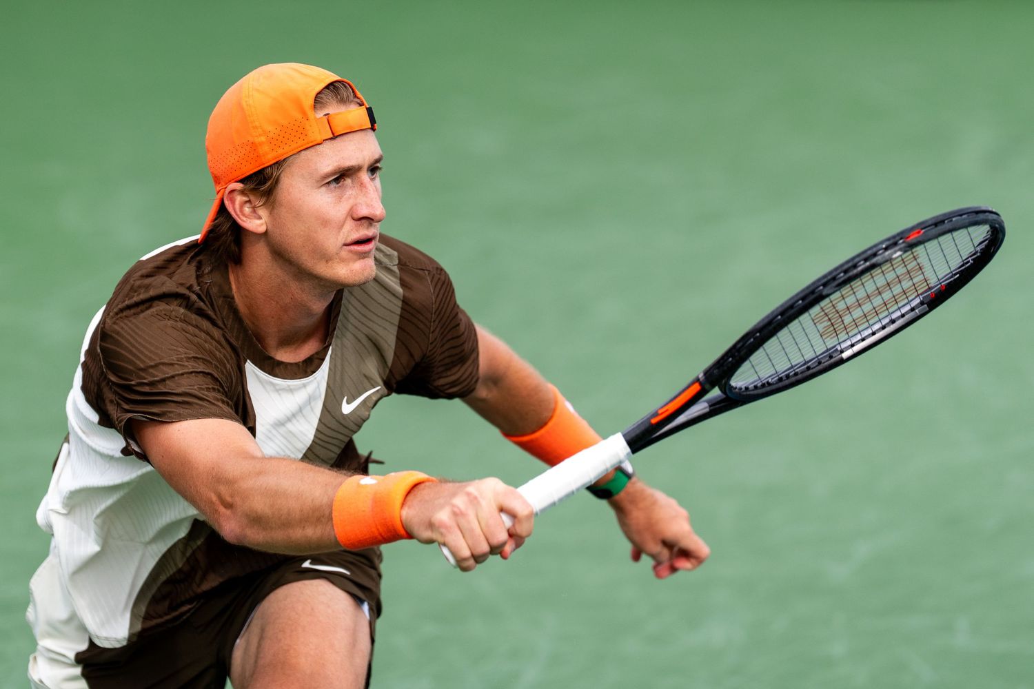 Male tennis player in brown Nike shirt and orange cap preparing to hit a backhand.