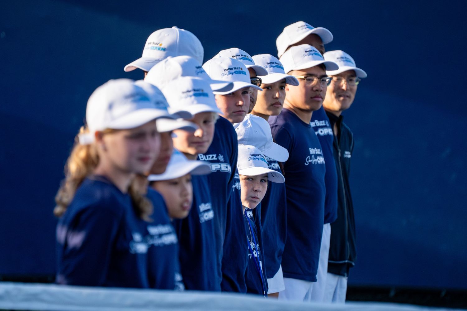 Group of young tennis ball kids in navy uniforms and white caps standing courtside.