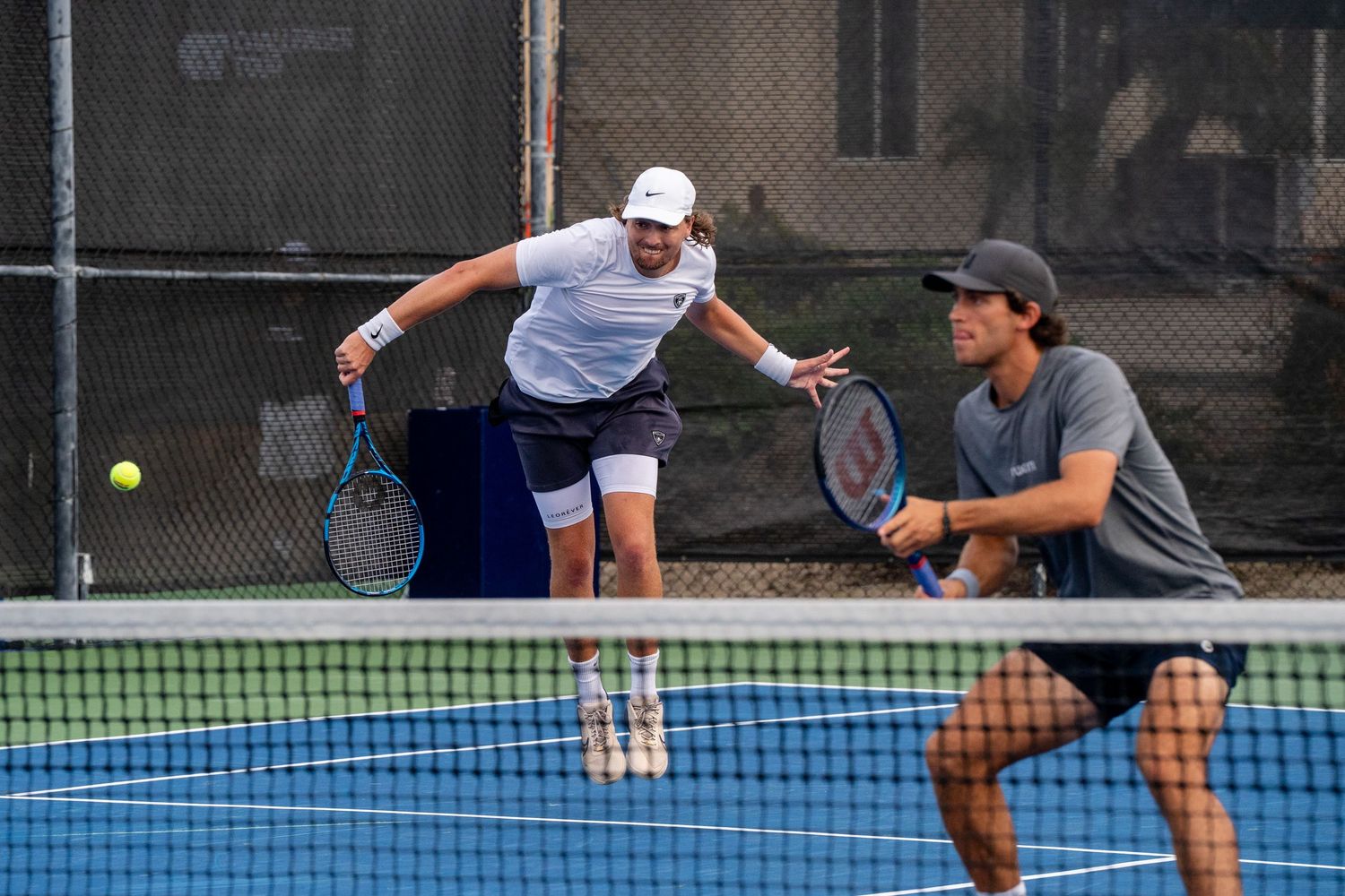 Two male tennis players compete in a doubles match on an outdoor hard court.