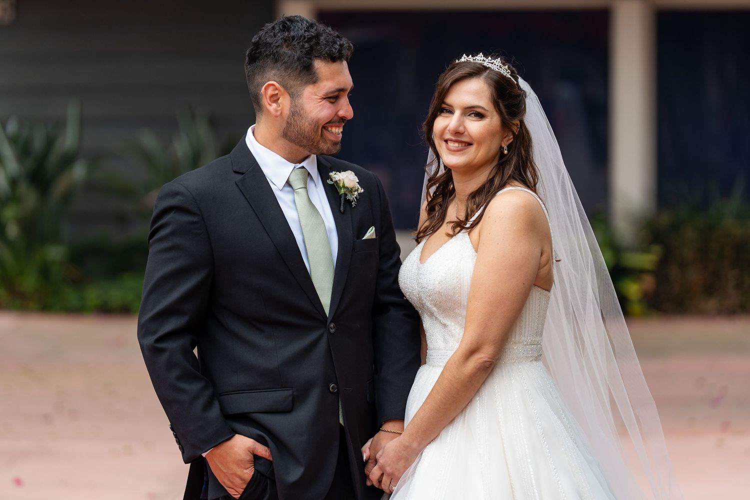 A smiling bride in a white gown and veil holds hands with a groom in a dark suit at an outdoor wedding.