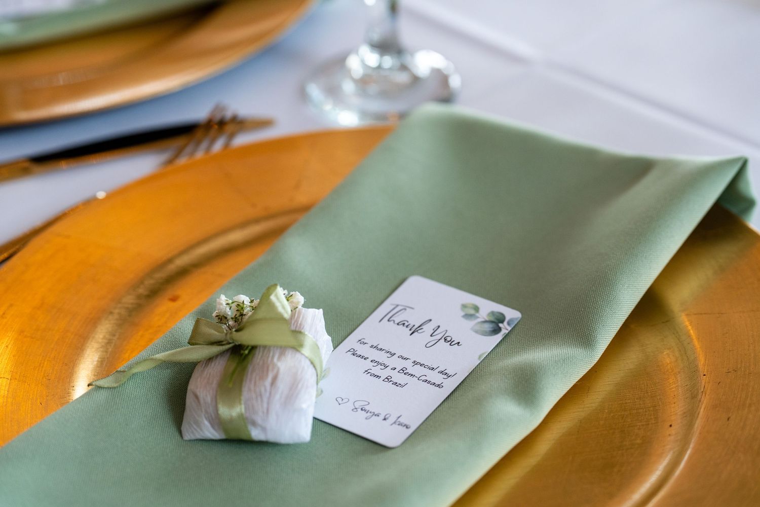 Elegant wedding table setting with sage green napkin, gold charger plate, and a small gift with thank you card.