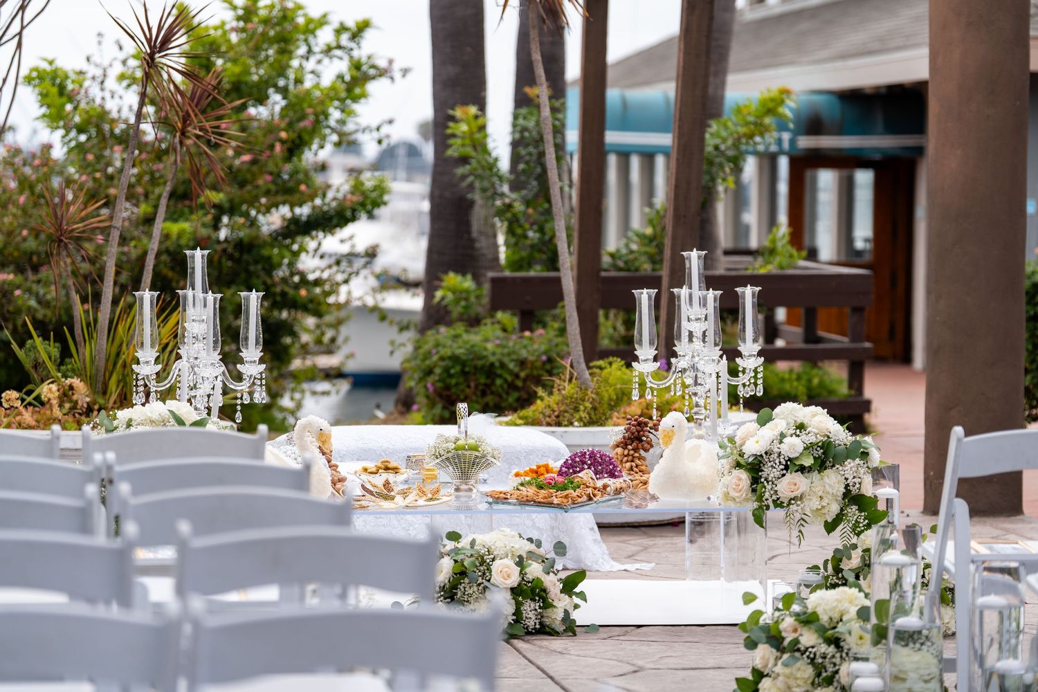 Elegant outdoor wedding reception with white chairs, floral arrangements, crystal candelabras, and a beautifully decorated buffet table.