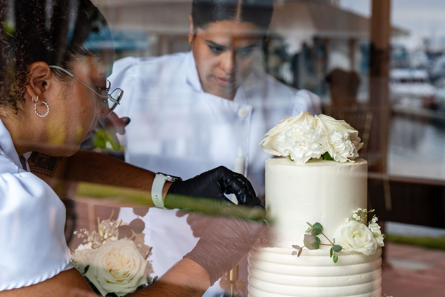 Two bakers decorating a white wedding cake with roses, reflected in glass window.