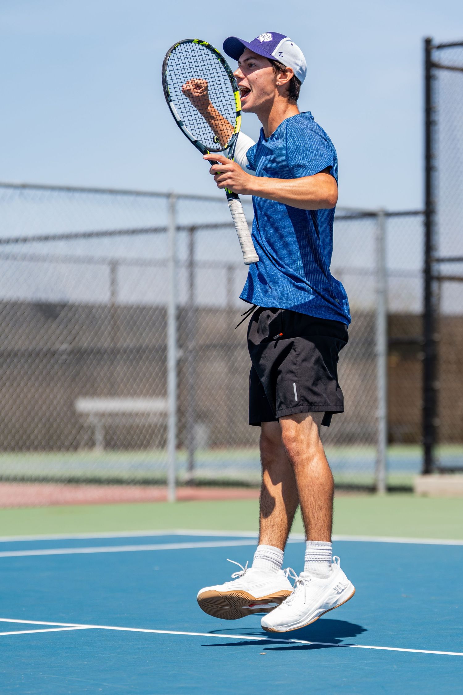 Excited male tennis player in blue shirt jumps and celebrates a point on outdoor court.