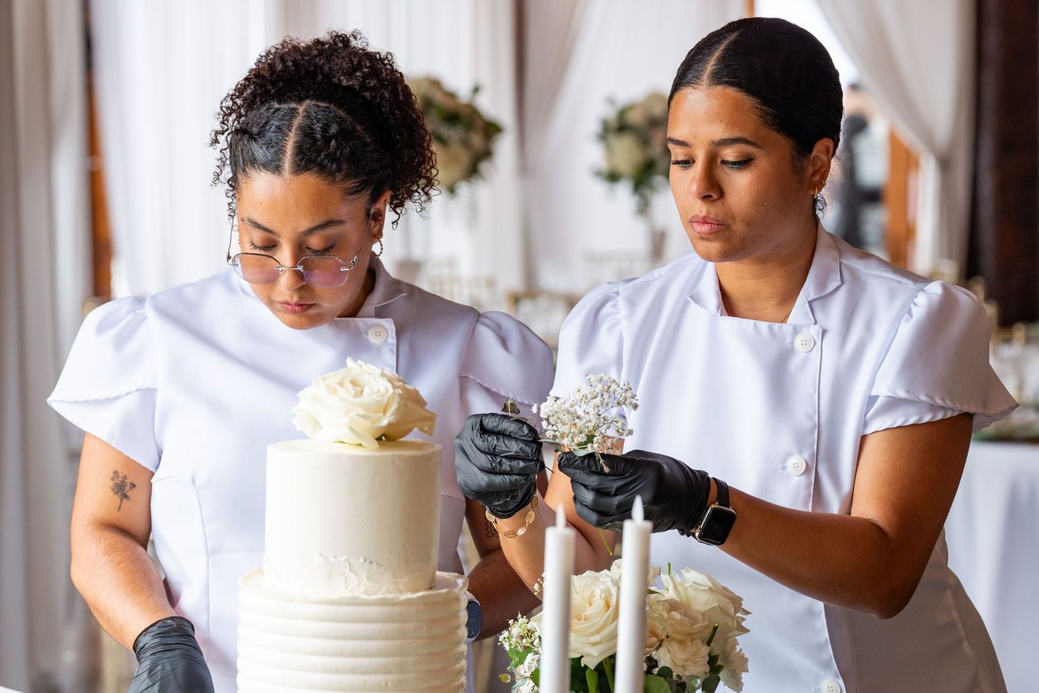 Two pastry chefs in white uniforms and black gloves decorating a elegant white three-tier wedding cake with flowers.