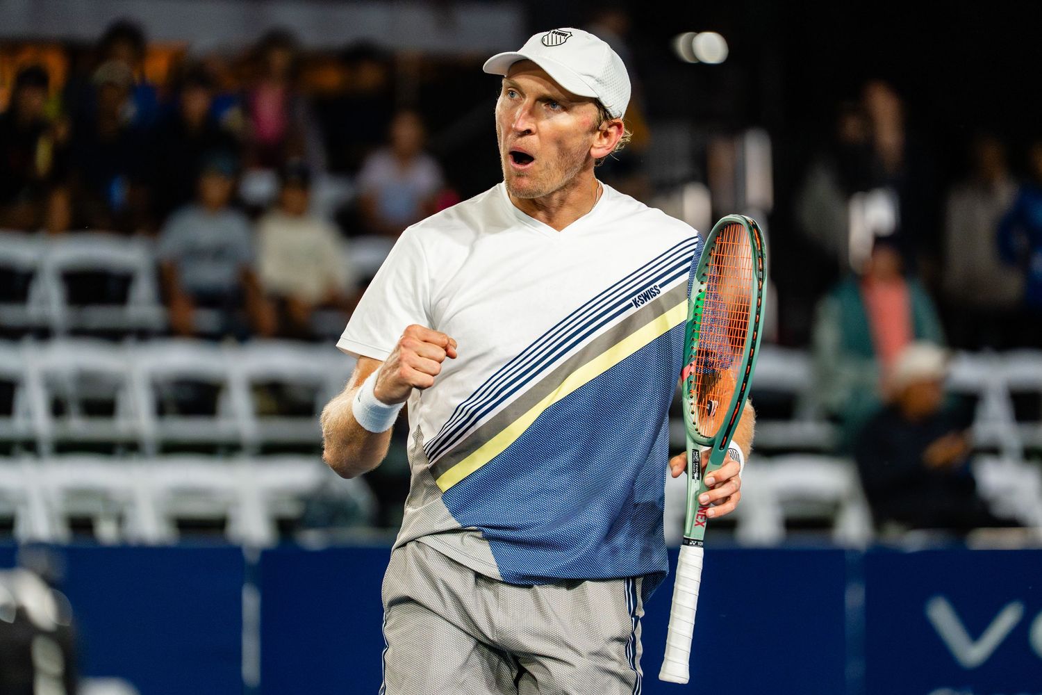 Tennis player celebrating on court, pumping fist while holding racket, wearing white cap and blue jersey.