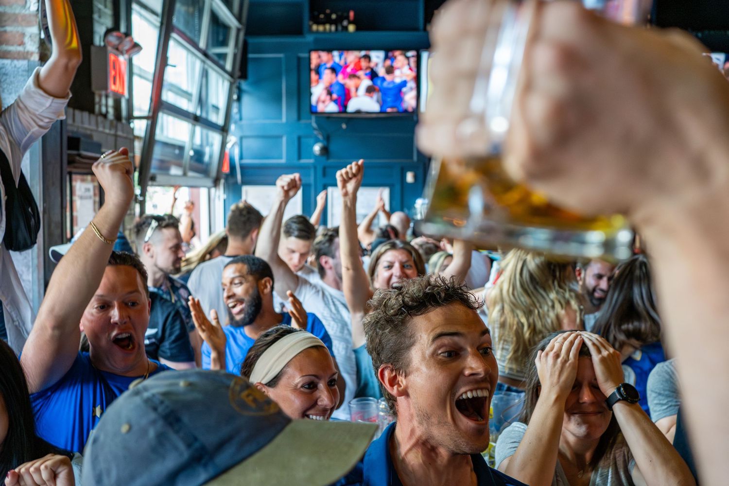 Excited sports fans celebrating at a bar, raising beers and cheering while watching a game on TV.