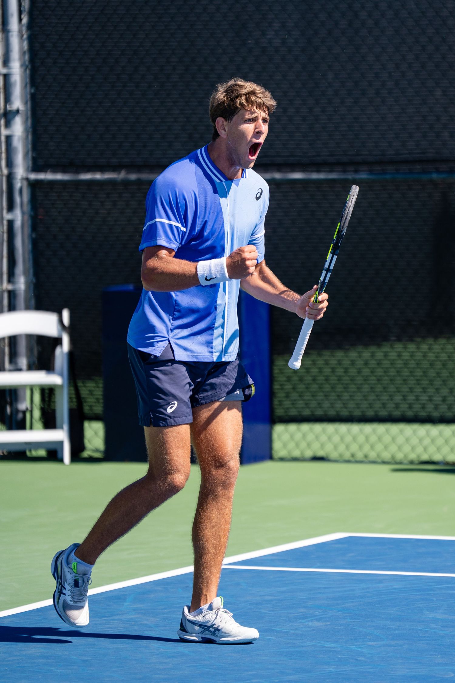 Young male tennis player in blue ASICS kit celebrates with a fist pump during an outdoor match.