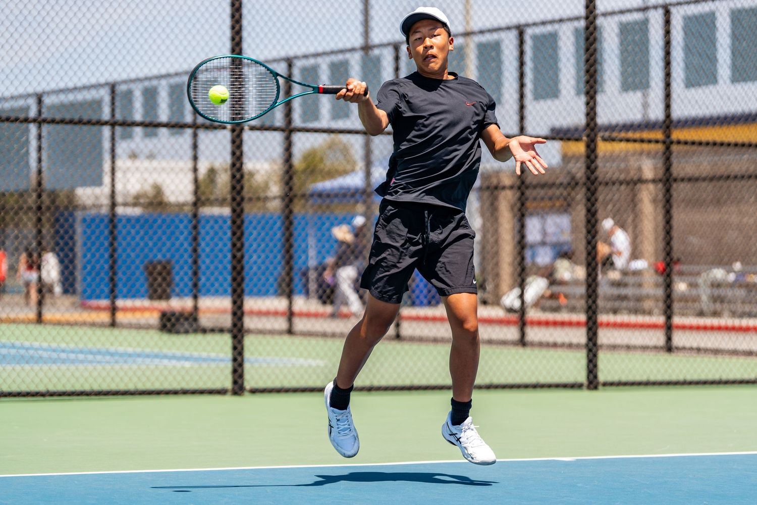 Young male tennis player leaps to hit a forehand shot on an outdoor blue tennis court.