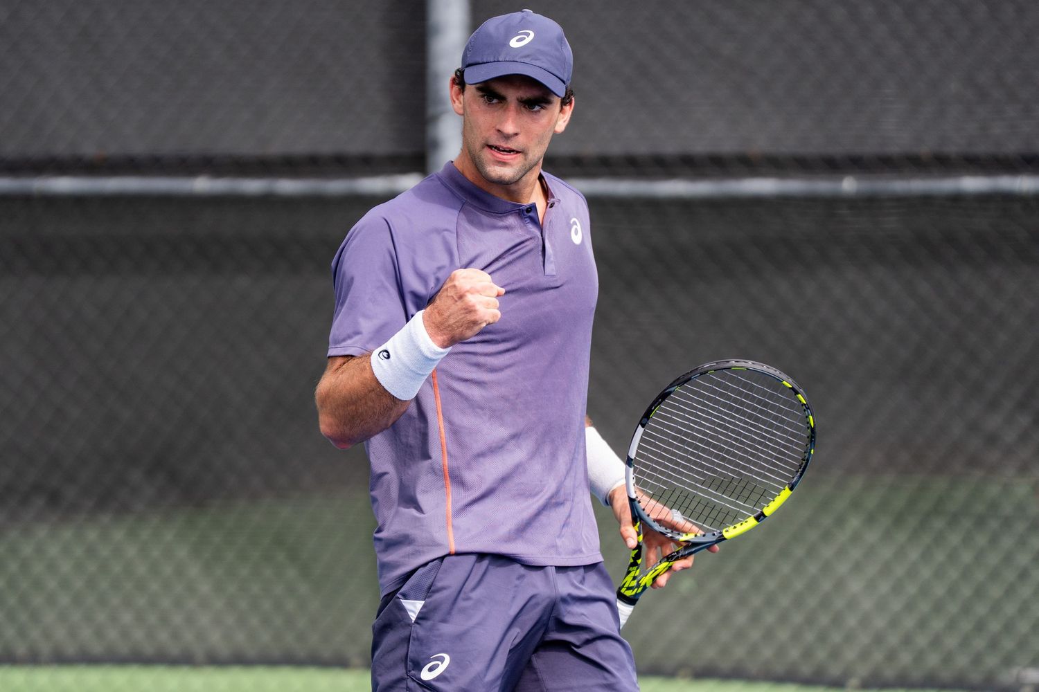 Male tennis player in navy Asics outfit celebrates with fist pump on court.