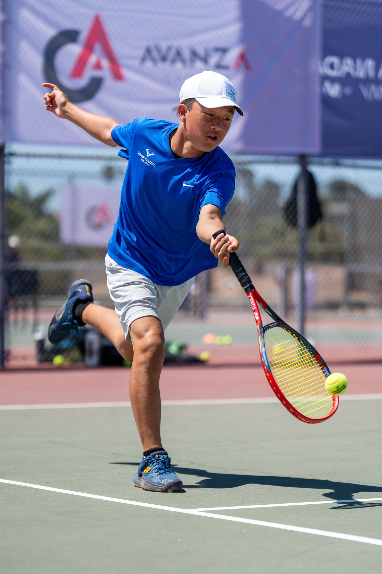Young tennis player in blue Nike shirt and white cap hitting a forehand shot on an outdoor court at an Avanza event.