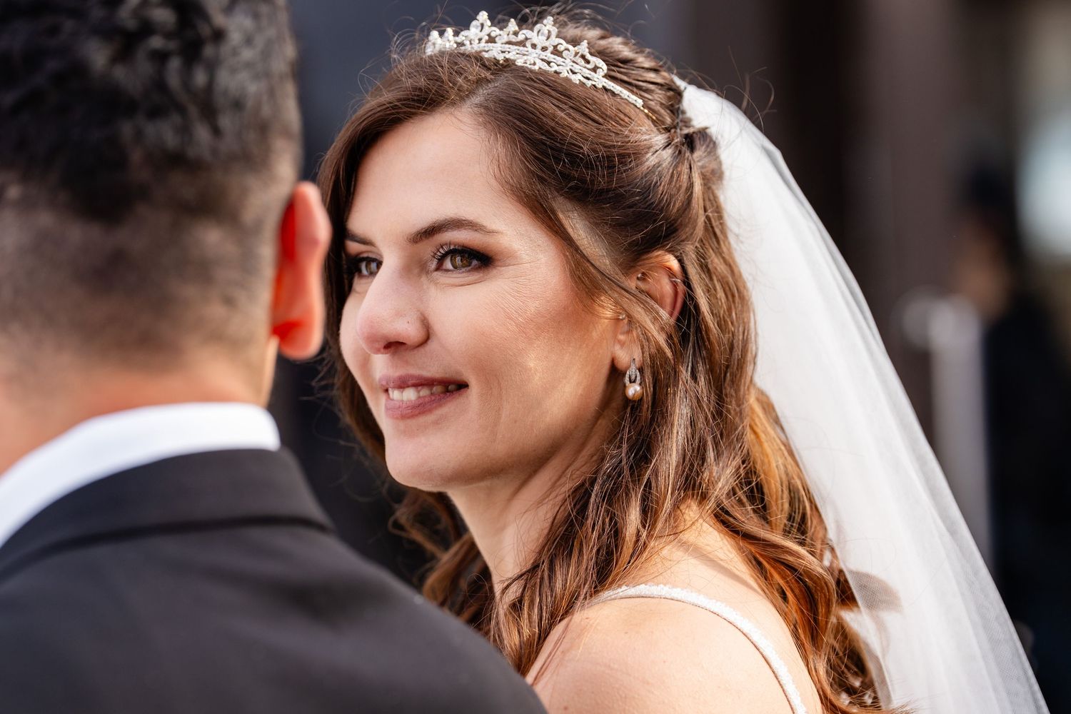 Bride wearing a tiara and veil smiles warmly at her groom during a wedding ceremony.