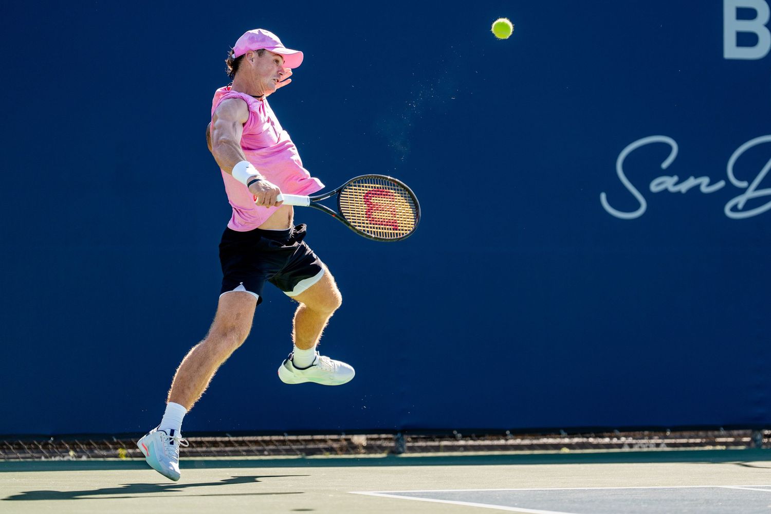 Tennis player in pink shirt leaping to hit a forehand shot at an outdoor ATP Challenger tournament.