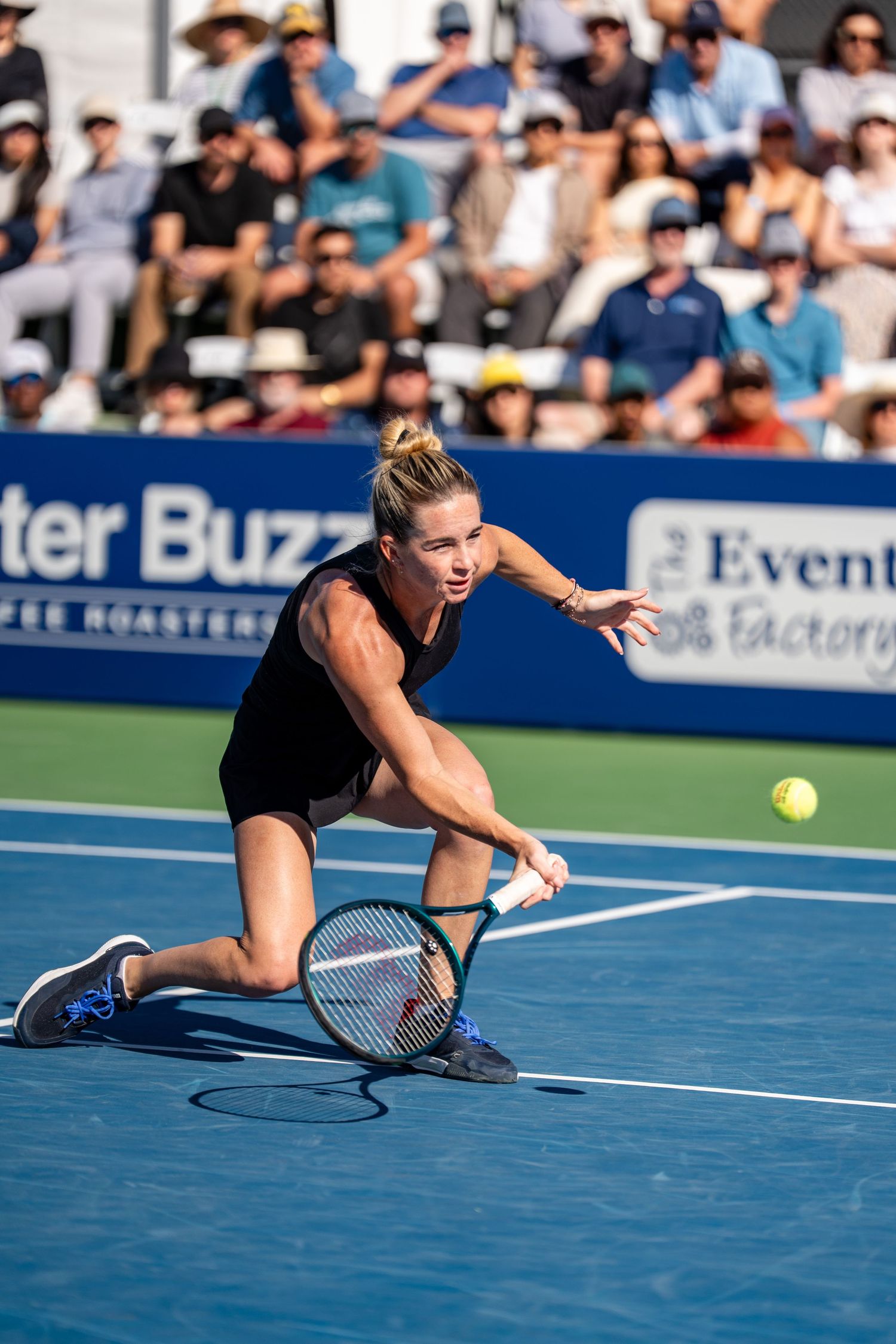 Female tennis player in black outfit lunging low to hit a forehand shot during a professional tennis match.