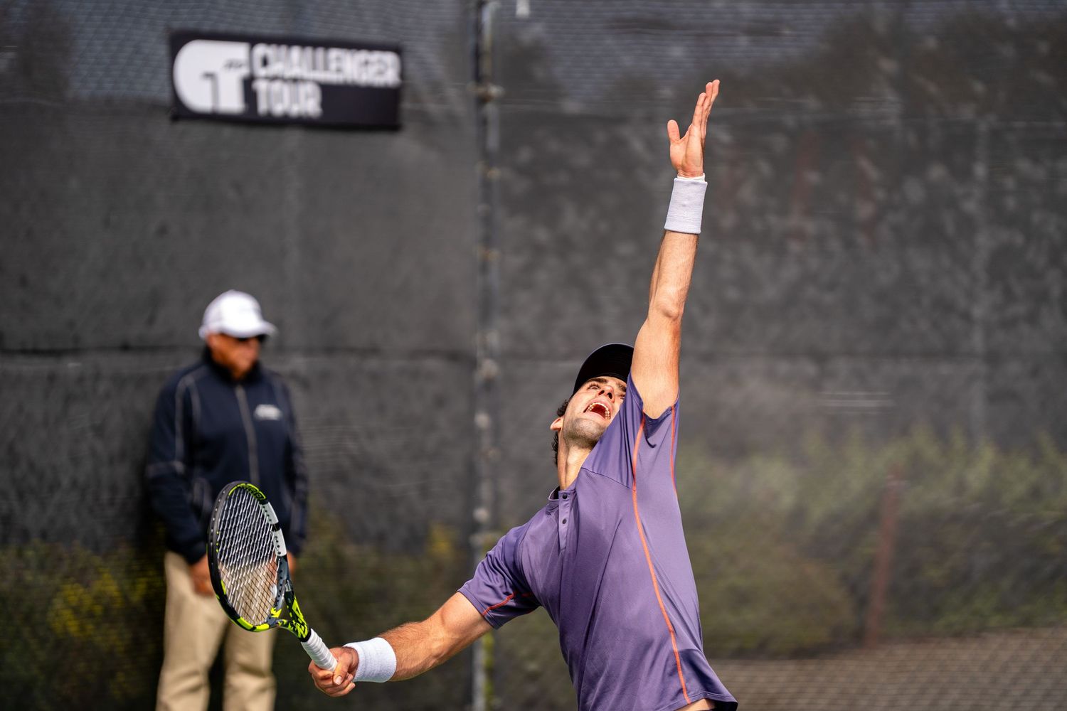 Tennis player in purple outfit tosses the ball high during a serve on an outdoor Challenger Tour court.
