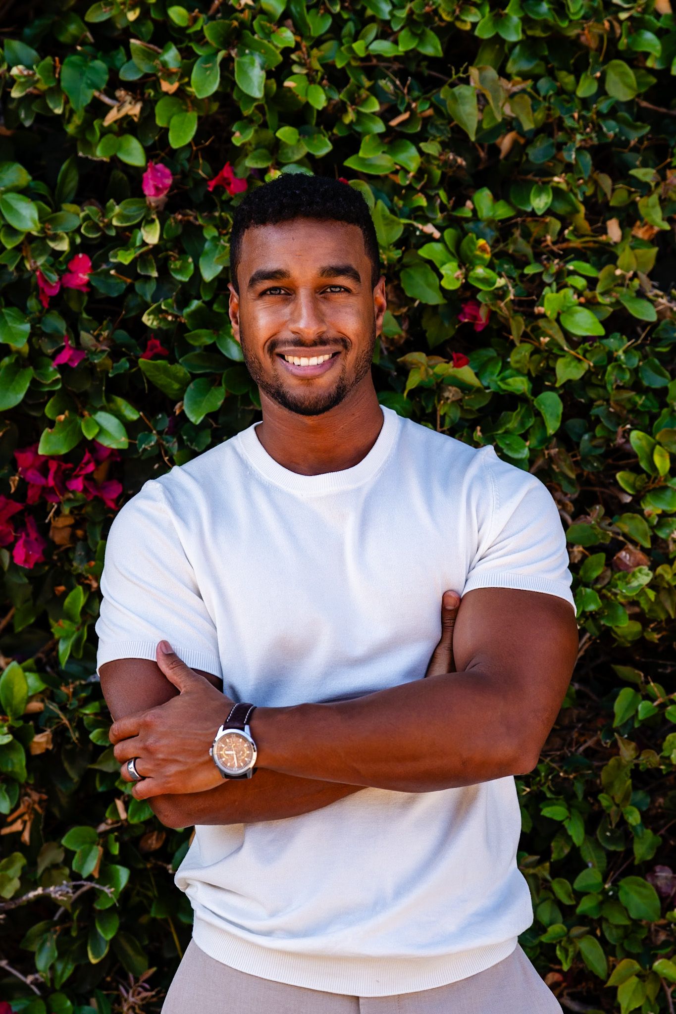 Smiling young Black man in white t-shirt standing with arms crossed in front of flowering green hedge.