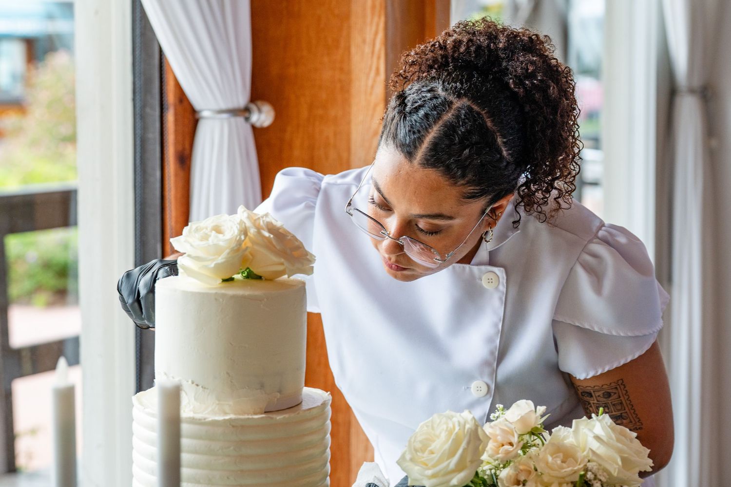 A baker in a white chef coat carefully decorates a elegant white tiered wedding cake with fresh roses.