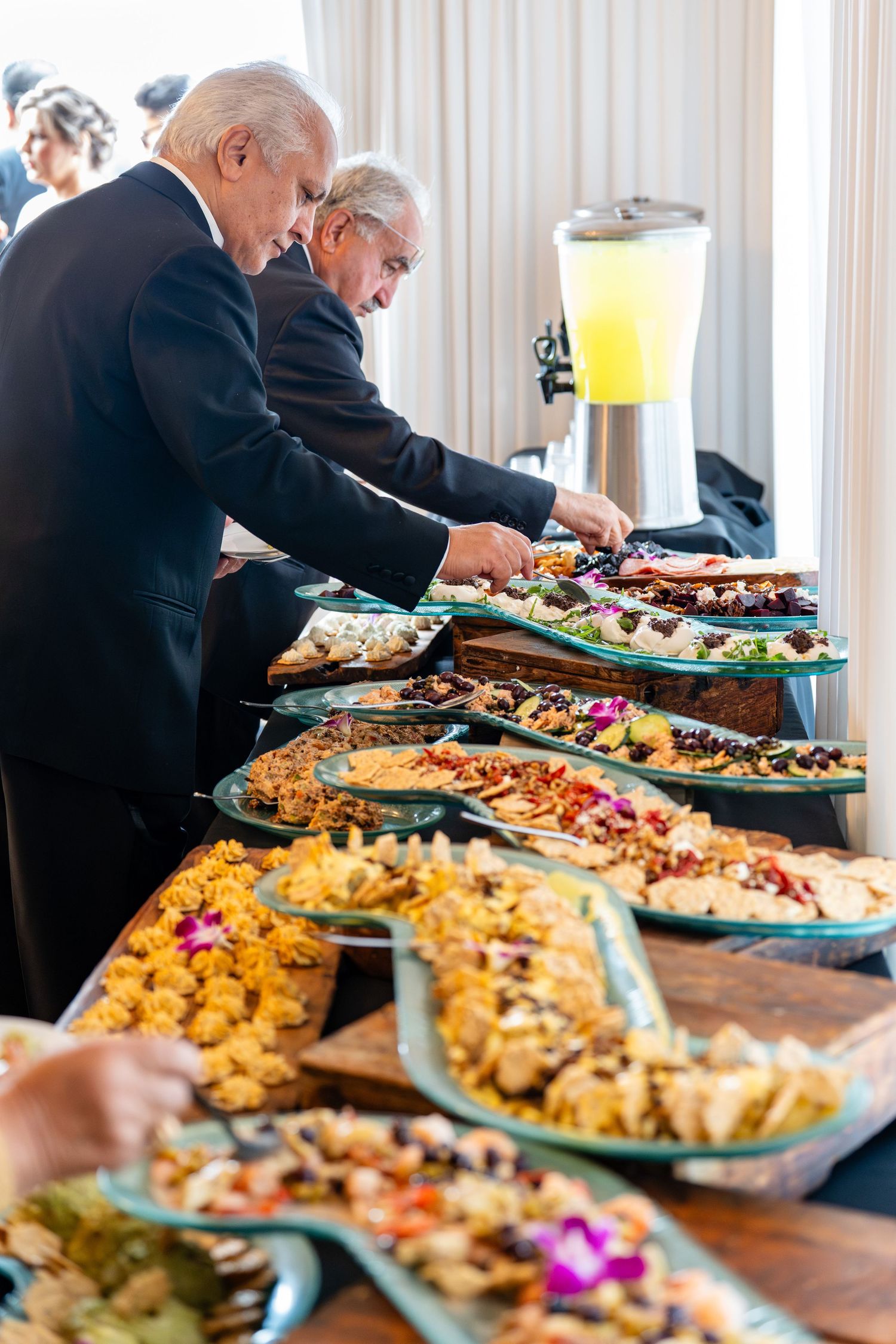 Two elegantly dressed men serving themselves at a lavish buffet table filled with colorful gourmet dishes at a formal event.