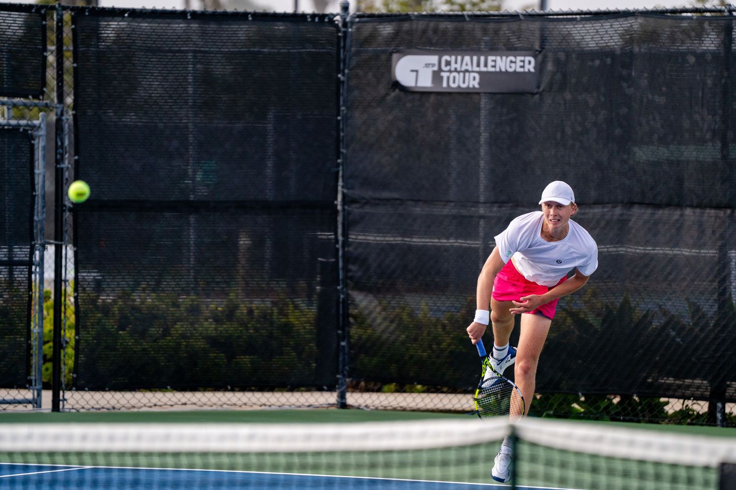 Tennis player in pink shorts serves on Challenger Tour outdoor court.