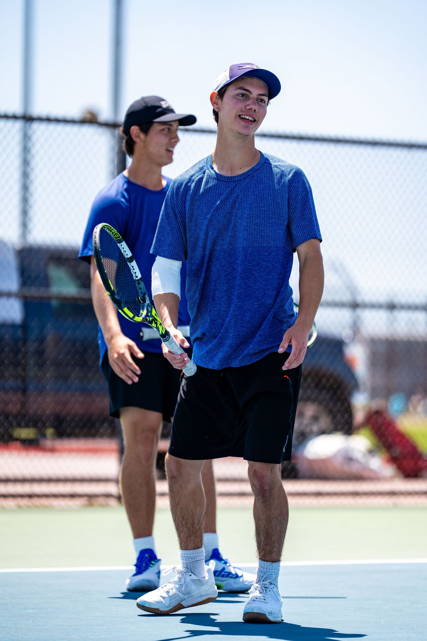 Two young male tennis players in blue shirts stand together on an outdoor tennis court.