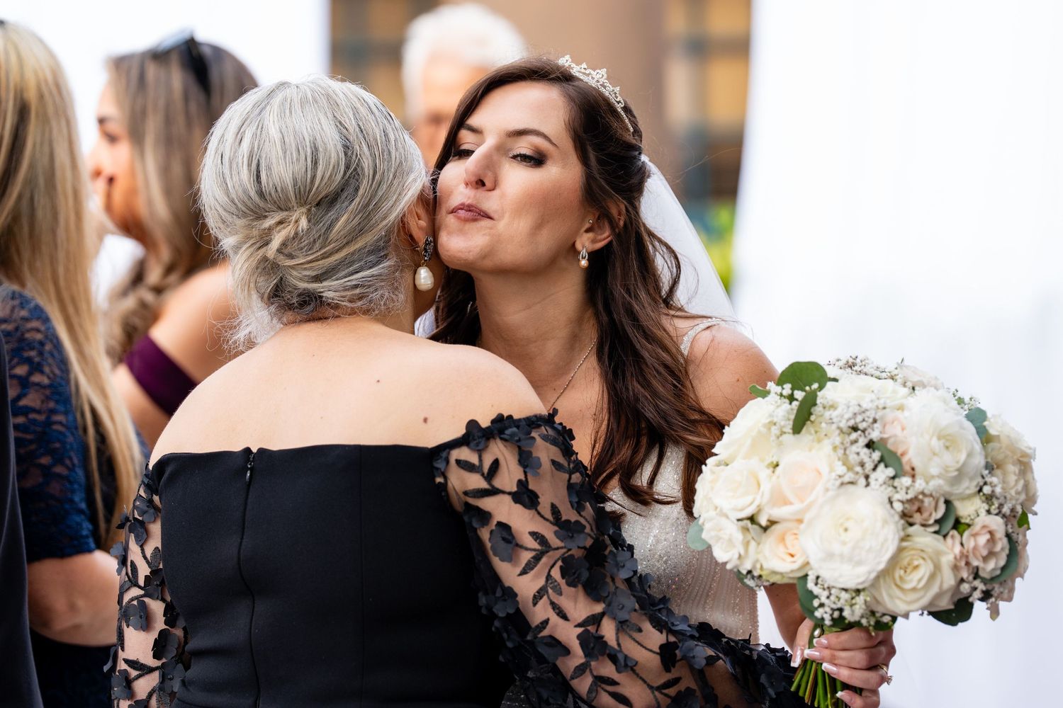 Bride in white gown hugging guest in black lace dress while holding white floral bouquet at wedding reception.
