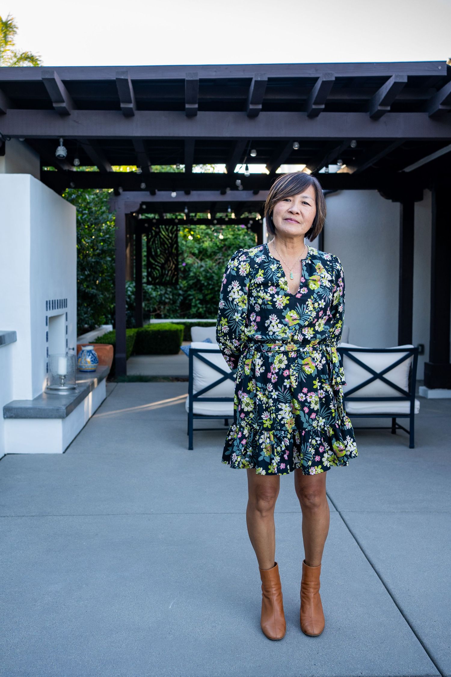 Asian woman in floral dress and brown boots standing on patio under pergola at dusk.