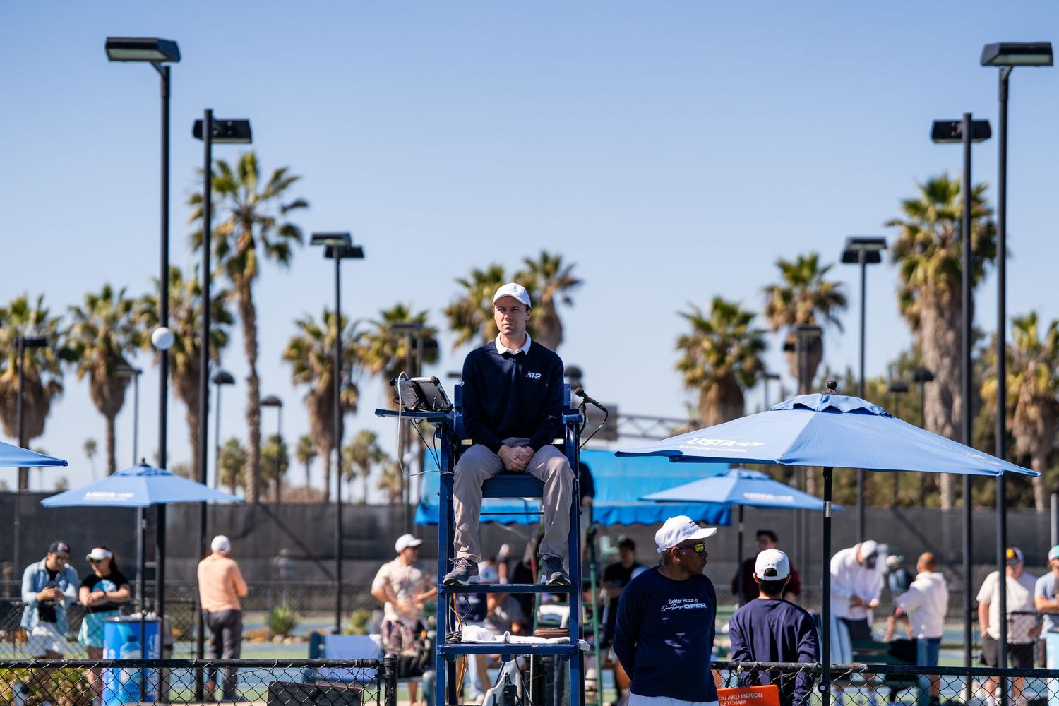 Chair umpire oversees match at outdoor tennis court with palm trees in background.