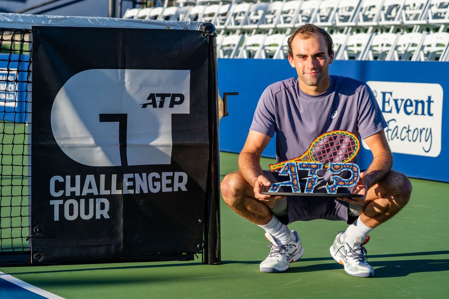 Tennis player poses with racket beside ATP Challenger Tour sign on court.