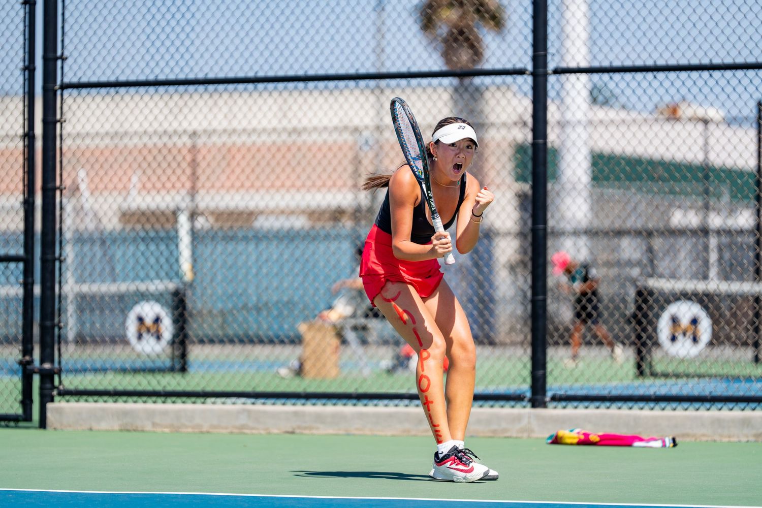 Female tennis player in red outfit prepares to serve on a sunny outdoor tennis court.
