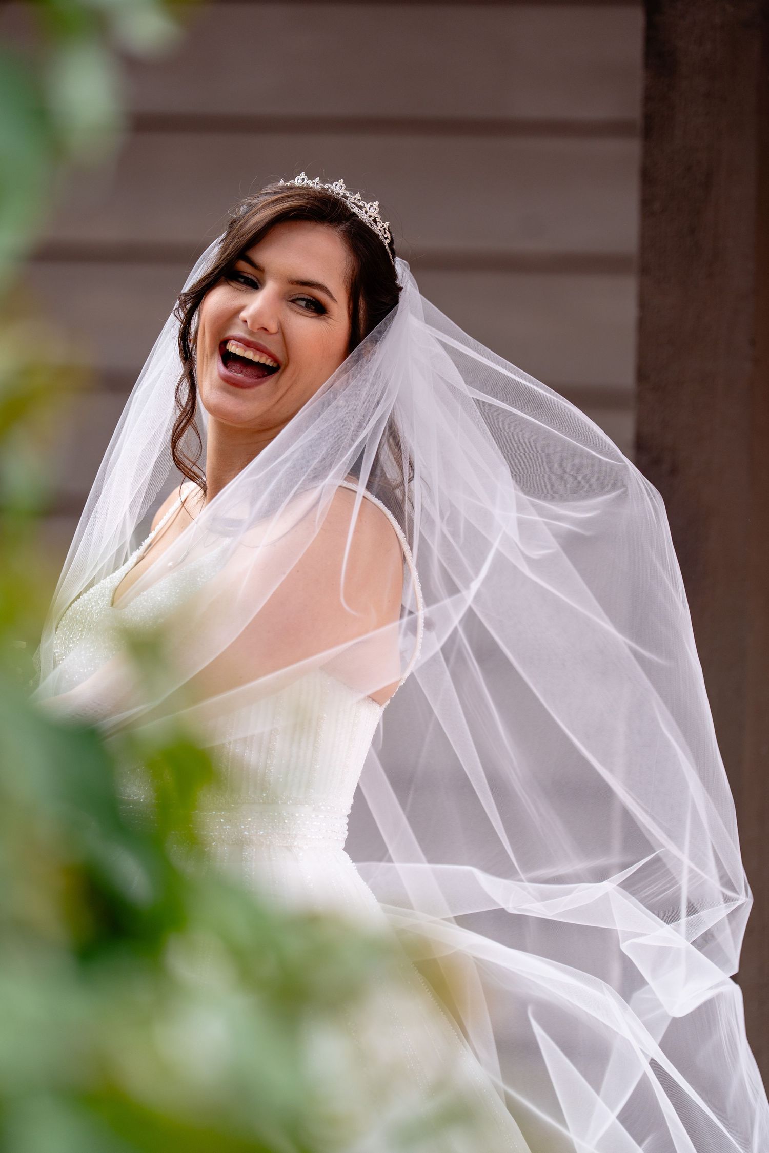Joyful bride in white gown, tiara, and flowing veil laughing outdoors on her wedding day.