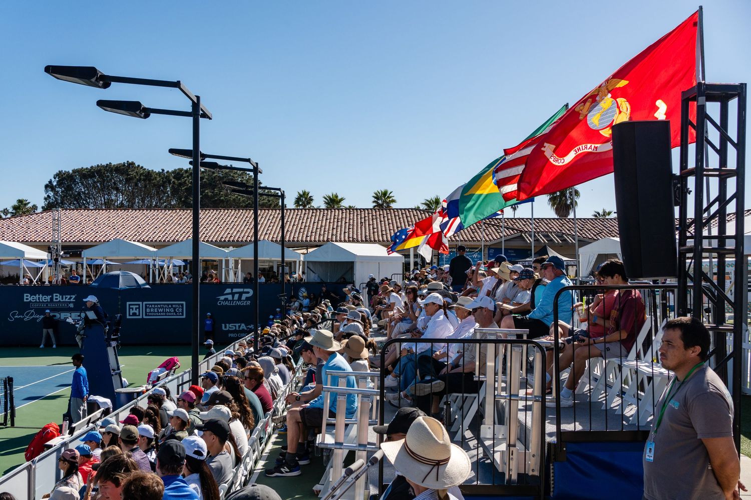 Crowd of spectators watches tennis matches under international flags at the San Diego Open ATP tournament.