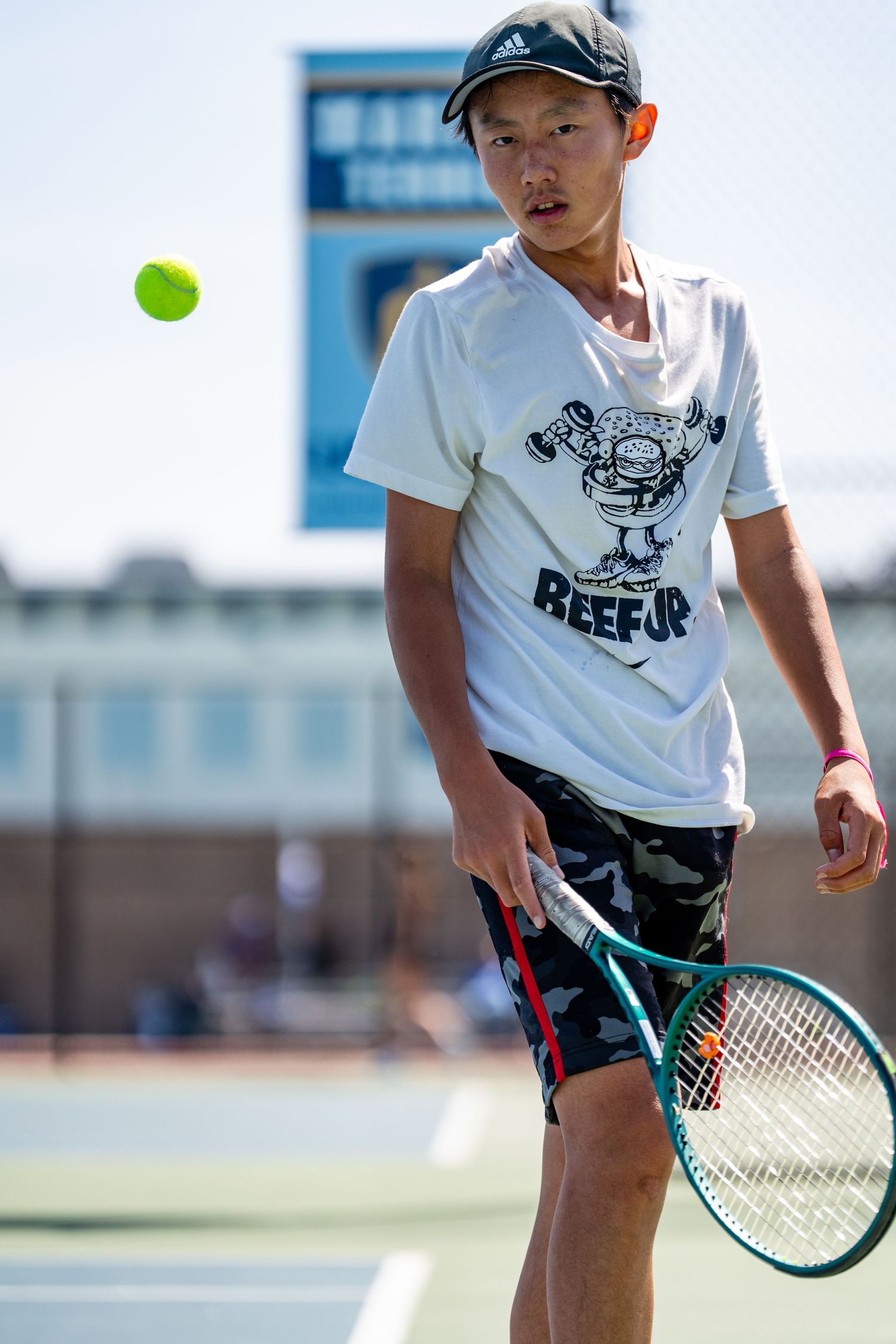 Young Asian male tennis player watches a tennis ball while preparing to serve on an outdoor hard court.