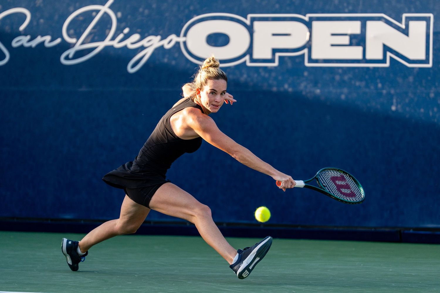 Female tennis player in black dress stretches for a backhand shot at the San Diego Open tournament.