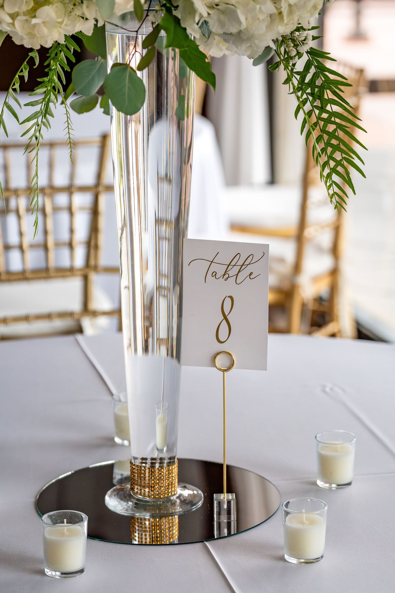 Elegant wedding table centerpiece with tall silver vase, white flowers, greenery, gold table number 8 sign, and votive candles.