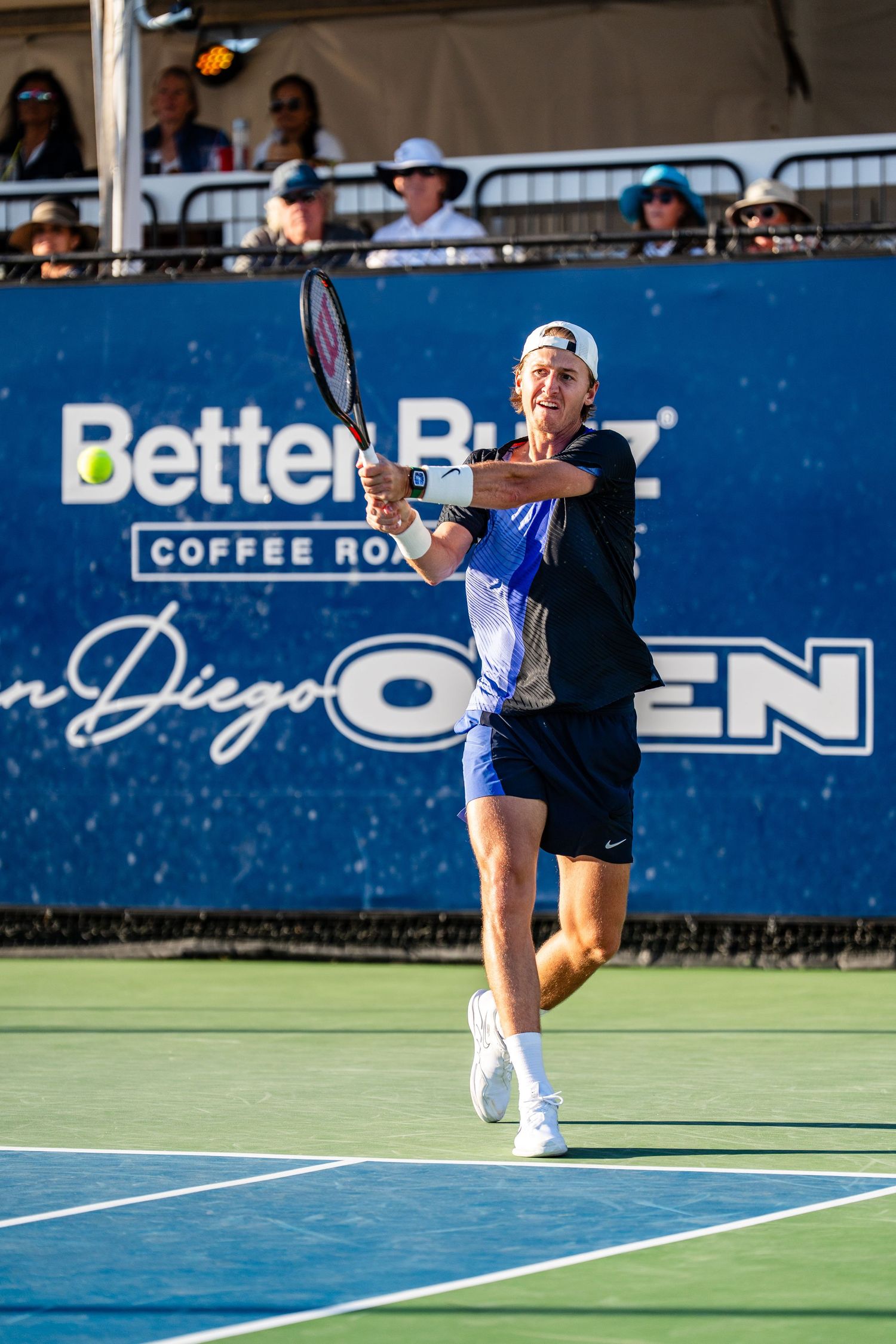 Tennis player hits a backhand shot at the San Diego Open with Better Buzz Coffee Roasters signage in background.