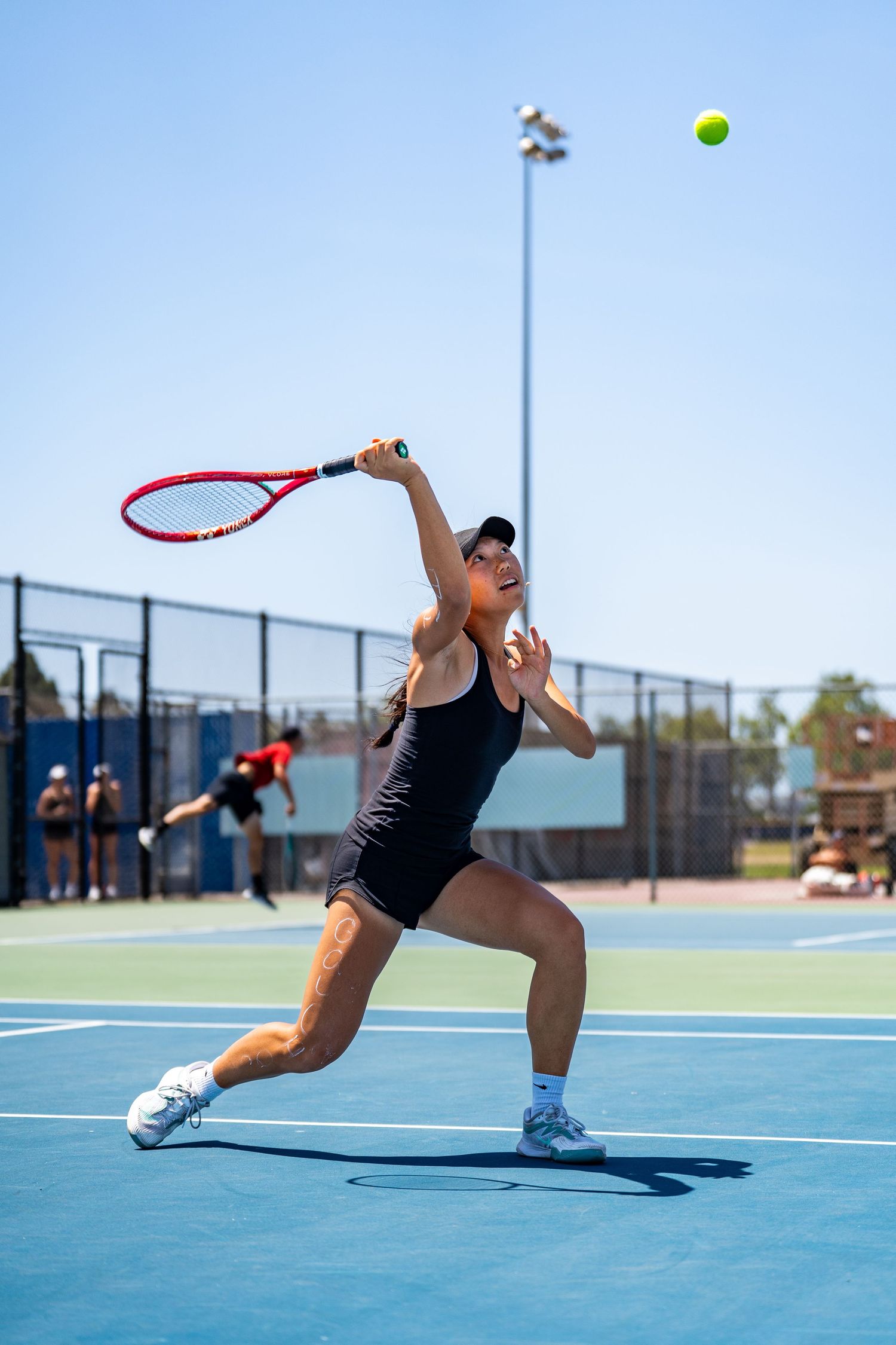 Female tennis player stretches to serve the ball on a sunny outdoor blue tennis court.