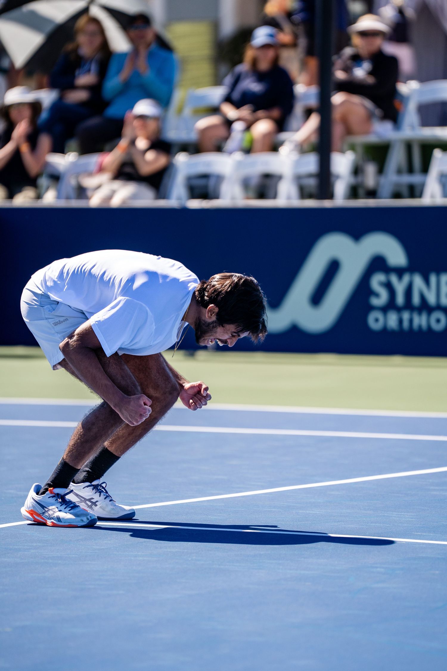 Tennis player bends forward in celebration or exhaustion on a blue hard court during a sunny match.