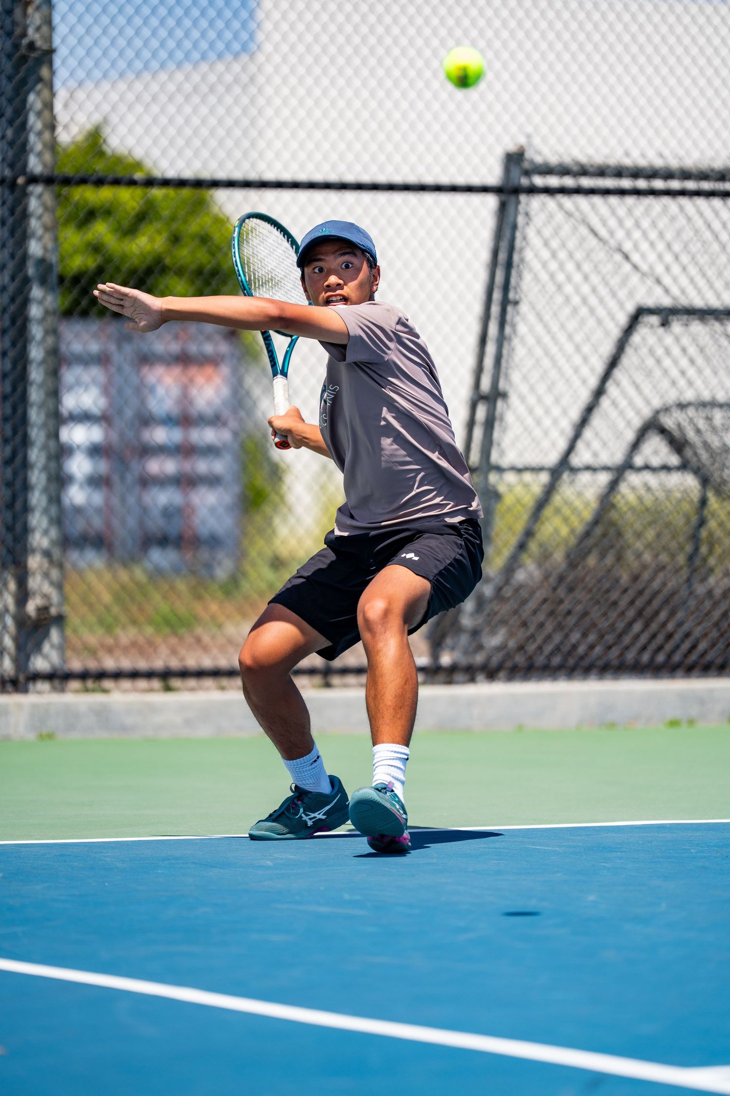 Male tennis player in gray shirt hitting a forehand on a bright blue outdoor court.