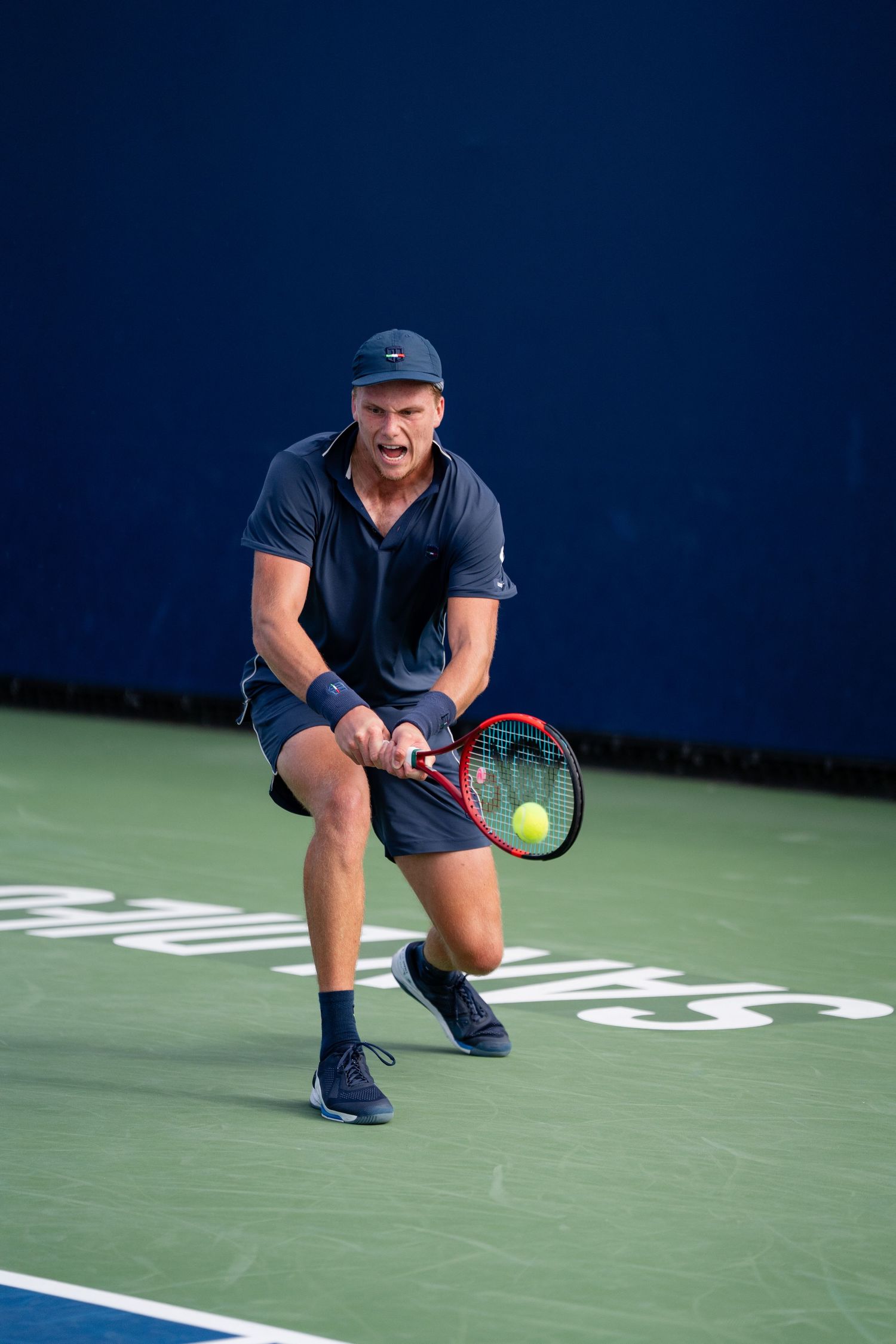 Male tennis player in all-navy outfit hitting a low backhand on a green hard court.